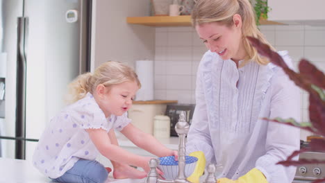 Mother-wearing-rubber-gloves-at-home-in-kitchen-with-young-daughter-having-fun-as-they-do-washing-up-at-sink--shot-in-slow-motion