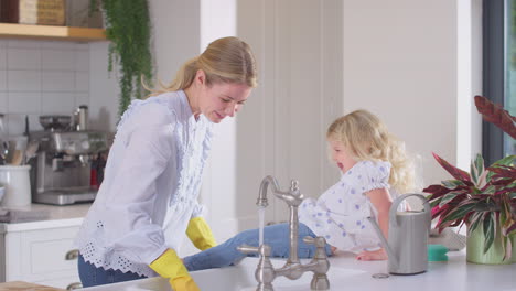 Mutter-Trägt-Gummihandschuhe-Zu-Hause-In-Der-Küche,-Während-Ihre-Kleine-Tochter-Spaß-Hat-Und-Mädchen-Die-Füße-Wäscht,-Während-Sie-Am-Waschbecken-Abwasch-Machen-–-Aufnahme-In-Zeitlupe