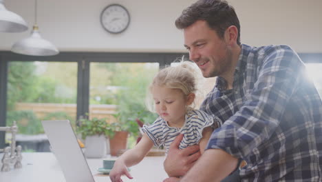 Working-father-using-laptop-at-home-on-kitchen-counter-whilst-looking-after-young-daughter---shot-in-slow-motion