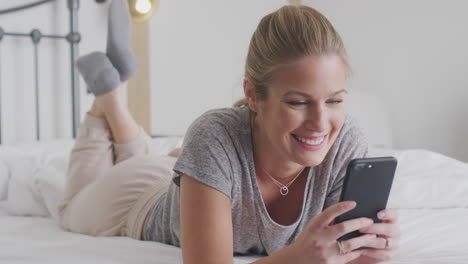 Smiling-Young-Woman-Lying-On-Bed-With-Mobile-Phone-Wearing-Pyjamas