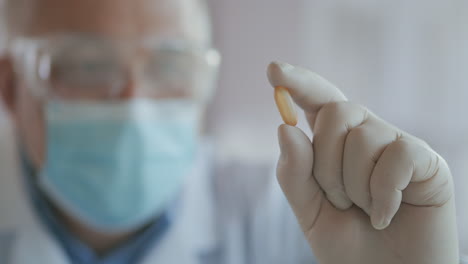 Extreme-Close-up-of-a-masked-scientist-developing-a-coronavirus-vaccine-holding-a-yellow-pill.-The-doctor-looks-at-the-painkillers-antiviral-medication.-Vitamins.-High-quality-4k-footage