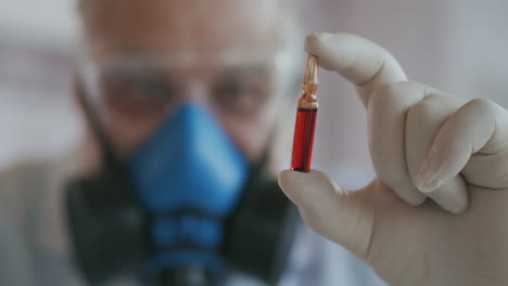 Extreme-Close-up-of-a-scientist-in-a-blue-respirator-and-protective-glasses-a-developer-of-a-coronavirus-vaccine-holding-a-red-ampoule.-The-doctor-looks-at-the-new-vaccine.-A-new-type-of-virus-medicine.-High-quality-4k-footage