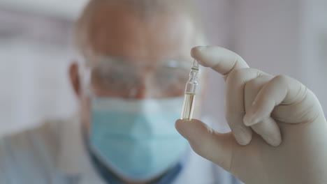 Close-up-a-masked-scientist-who-developed-a-coronavirus-vaccine-holds-an-ampoule-of-white-liquid-and-examines-the-vaccine.-The-doctor-looks-at-the-medicine-before-applying-it.-High-quality-4k-footage