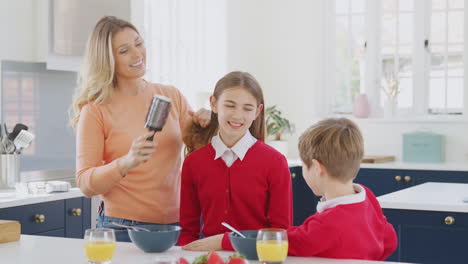 Mother-Brushes-Daughter's-Hair-As-Children-In-School-Uniform-Eat-Breakfast-At-Home