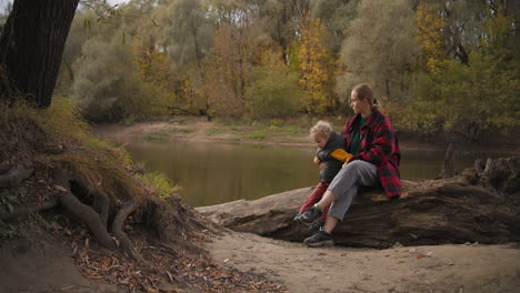 caucasian-woman-with-little-child-are-resting-on-coast-of-small-river-in-park-enjoying-good-fall-weather-happy-family-at-nature