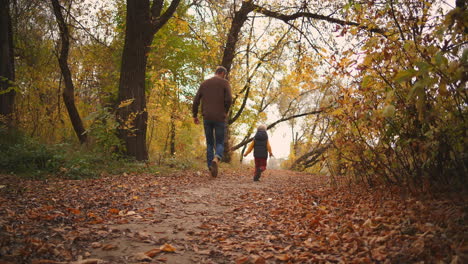Glückliches-Kind-Und-Vater-Im-Herbstlichen-Wald,-Der-über-Einen-Mit-Trockenem-Laub-Bedeckten-Weg-Rennt,-Mann-Fängt-Sohn-Und-Wirbelt-Glückliches-Familienwochenende-In-Der-Natur