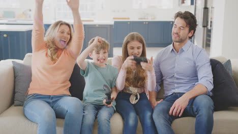Parents-Having-Fun-Sitting-On-Sofa-With-Children-At-Home-Playing-On-Games-Console