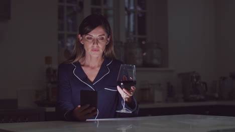 Unglückliche-Frau-Im-Schlafanzug-Sitzt-Nachts-Mit-Einem-Glas-Wein-In-Der-Küche-Und-Benutzt-Ihr-Mobiltelefon