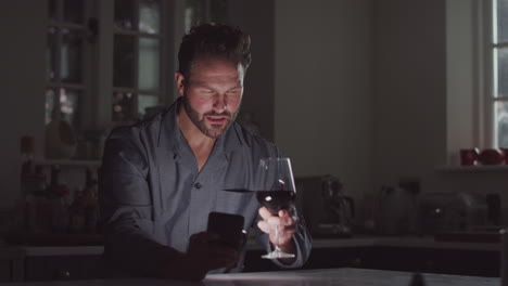 Mature-Man-Wearing-Pyjamas-Sitting-In-Kitchen-With-Glass-Of-Wine-At-Night-Using-Mobile-Phone