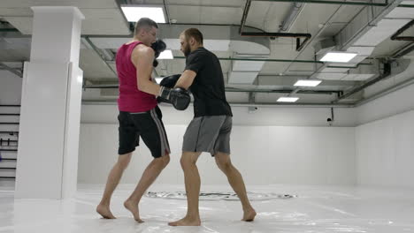 Two-men-in-a-white-gym.-The-coach-and-the-fighter-work-out-the-kick-in-the-jump-knee-on-the-paws-in-slow-motion