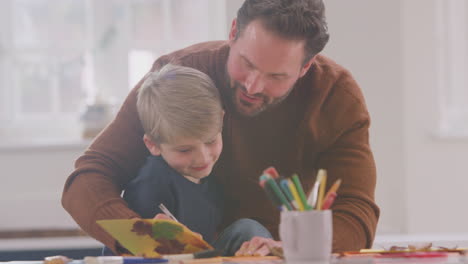 Father-With-Son-At-Home-Doing-Craft-And-Making-Picture-From-Leaves-In-Kitchen