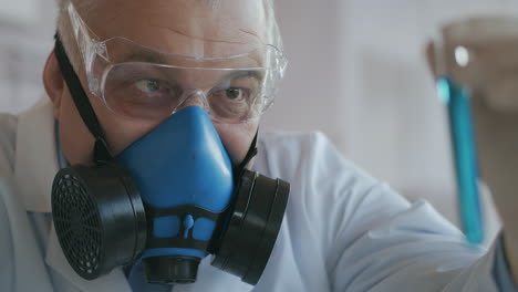 A-scientist-at-a-pharmaceutical-company-developing-drugs-wearing-a-blue-respirator-and-safety-glasses-holds-a-new-medicine-a-glass-tube-with-a-blue-liquid-in-his-hands-and-looks-at-it.-High-quality-4k-footage