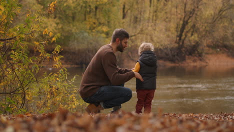 little-child-is-giving-five-to-father-and-laughing-resting-together-at-forest-with-picturesque-lake-at-autumn-day-family-trip-and-communication