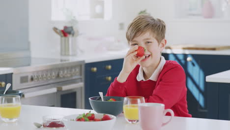 Lachender-Junge-In-Schuluniform-In-Der-Küche,-Der-Beim-Frühstück-Erdbeeren-Auf-Die-Nase-Setzt