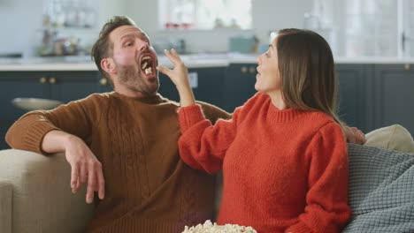 Man-Catching-Popcorn-In-Mouth-As-Woman-Throws-It-To-Him-Sitting-On-Sofa