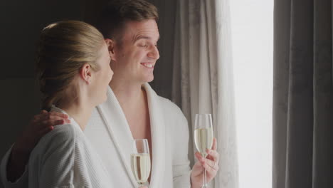 Couple-Wearing-Bathrobes-On-Romantic-Hotel-Break-Standing-By-Open-Curtains-With-Glasses-Of-Champagne