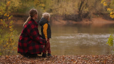 family-rest-in-autumn-forest-mother-and-little-son-are-viewing-lake-standing-on-coast-with-dry-grass-and-foliage-yellowed-trees-around