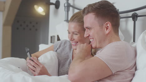 Couple-In-Bed-Wearing-Pyjamas-Looking-At-Mobile-Phone-Together