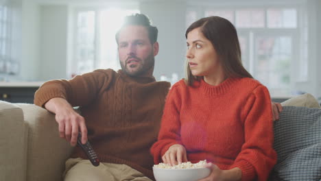 Couple-Sitting-On-Sofa-Spilling-Popcorn-As-They-Watch-Thriller-On-TV-Together