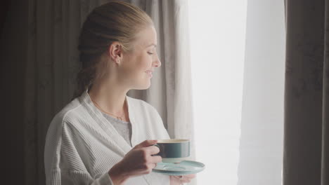 Woman-Wearing-Bathrobe-On-Hotel-Or-Spa-Break-Standing-By-Open-Curtains-With-Hot-Drink