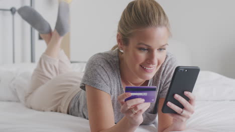 Frau-Liegt-Mit-Mobiltelefon-Im-Bett-Und-Kauft-Online-Mit-Kreditkarte-Ein-Und-Trägt-Pyjamas