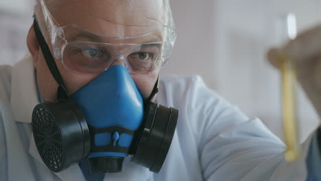 A-scientist-at-a-pharmaceutical-company-developing-drugs-wearing-a-blue-respirator-and-safety-glasses-holds-a-new-drug-a-glass-tube-with-a-yellow-liquid-in-his-hands-and-looks-at-it.-High-quality-4k-footage