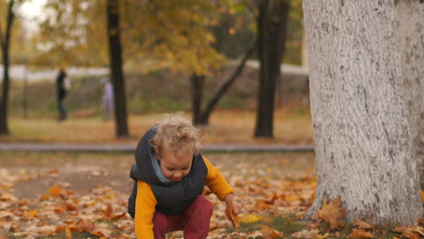 Der-Glückliche-Kleine-Junge-Spielt-Am-Herbsttag-Im-Park-Mit-Gelben,-Trockenen-Blättern,-Wirft-Laub-Und-Lächelt-Und-Hat-Spaß-Beim-Spazierengehen