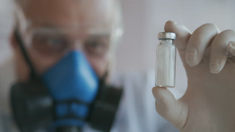 Extreme-Close-up-of-a-scientist-in-a-blue-respirator-and-protective-glasses-who-developed-a-coronavirus-vaccine-holding-an-ampoule-of-white-powder.-Narcotic-substances-and-painkillers.-High-quality-4k-footage