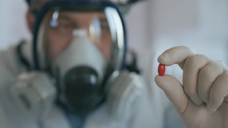 Close-up-in-the-laboratory-a-man-in-a-glass-respirator-screen-holds-a-new-medicine-a-red-pill-an-antibiotic-against-the-virus-and-looks-at-it.-High-quality-4k-footage