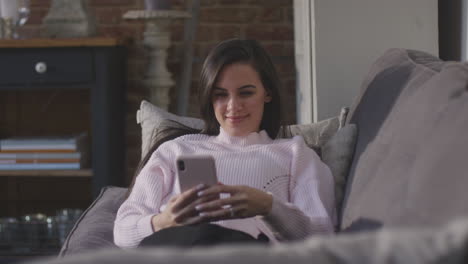 Smiling-Woman-Relaxing-On-Sofa-At-Home-Checking-Social-Media-On-Mobile-Phone