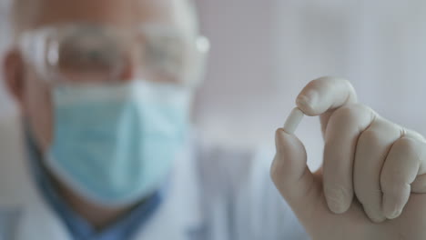 Extreme-Close-up-of-a-masked-scientist-developing-a-coronavirus-vaccine-holding-a-white-pill.-The-doctor-looks-at-the-painkillers-antiviral-medication.-Vitamins.-High-quality-4k-footage