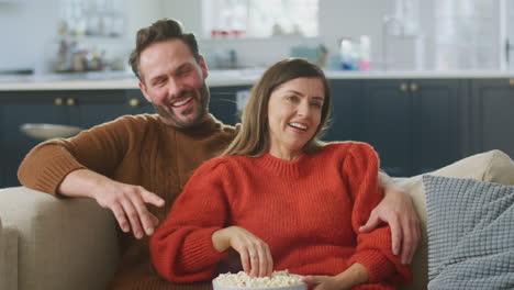 Couple-Sitting-On-Sofa-With-Popcorn-Laughing-Watching-Comedy-On-TV-Together