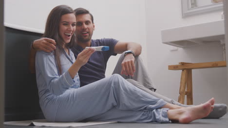 Excited-Couple-Sitting-On-Floor-In-Bathroom-At-Home-With-Positive-Home-Pregnancy-Test
