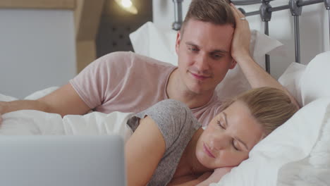 Couple-In-Bed-Wearing-Pyjamas-Streaming-Movie-On-Laptop-As-Woman-Sleeps
