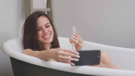 Woman-Lying-And-Relaxing-In-Bath-At-Home-Streaming-Movie-On-Mobile-Phone-And-Drinking-Glass-Of-Wine