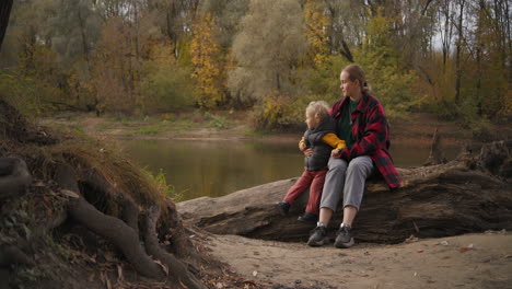 early-fall-in-forest-people-are-walking-at-good-weather-mother-and-child-are-sitting-on-shore-of-river-family-walk-at-nature