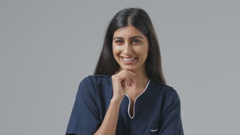 Studio-Portrait-Of-Smiling-Female-Nurse-In-Uniform-Resting-Chin-On-Hand-Against-Plain-Background