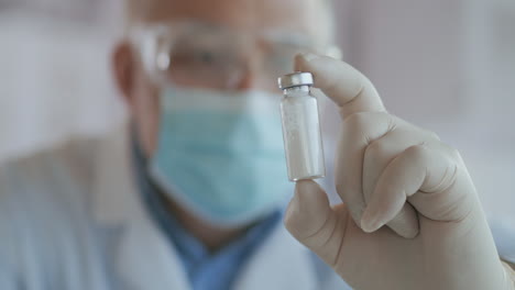 Close-up-a-masked-scientist-who-developed-a-coronavirus-vaccine-holds-an-ampoule-of-white-powder-and-examines-the-vaccine.-The-doctor-looks-at-the-antibiotics-before-use.-High-quality-4k-footage