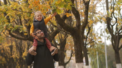 happy-son-and-father-are-walking-in-forest-at-autumn-baby-is-sitting-on-shoulders-of-dad-and-viewing-leaves-of-tree-joyful-weeknd-in-park