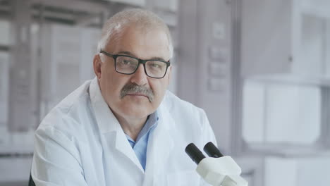 Portrait-a-white-haired-male-lab-doctor-with-a-microscope-looks-at-the-camera-serious-and-then-smiles-in-slow-motion.-High-quality-4k-footage