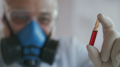 Extreme-Close-up-of-a-scientist-in-a-blue-respirator-and-protective-glasses-a-developer-of-a-coronavirus-vaccine-holding-a-red-ampoule.-The-doctor-looks-at-the-new-vaccine.-A-new-type-of-virus-medicine.-High-quality-4k-footage