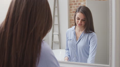 Woman-Wearing-Pyjamas-At-Home-In-Modern-Bathroom-Brushing-Teeth-Reflected-In-Mirror