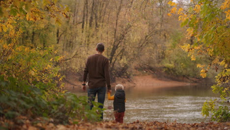 cute-toddler-is-holding-hand-of-his-father-in-forest-man-and-child-are-standing-together-on-calm-shore-of-lake-at-autumn-day-idyllic-picture-of-famly-rest-at-weekends