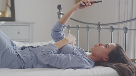 Woman-Wearing-Pyjamas-Posing-And-Taking-Selfies-On-Mobile-Phone-Lying-On-Bed