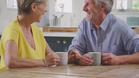 Kissing-Retired-Couple-Sitting-Around-Table-At-Home-Having-Morning-Coffee-Together