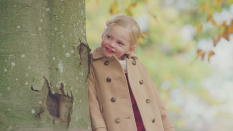 Young-Girl-Looking-Around-Trunk-Of-Autumn-Tree-Playing-Hide-And-Seek-In-Garden