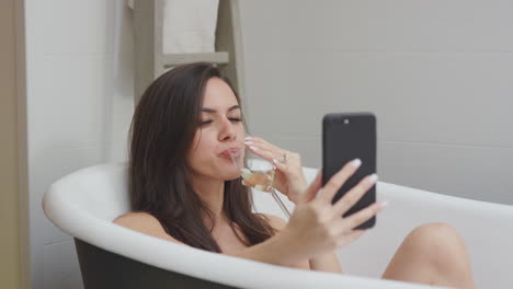 Woman-Lying-And-Relaxing-In-Bath-At-Home-Posing-For-Selfie-On-Mobile-Phone-And-Drinking-Glass-Of-Wine