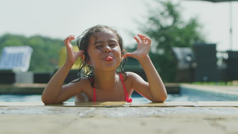 Portrait-Of-Young-Girl-Looking-Over-Edge-Of-Outdoor-Pool-Pulling-Funny-Face-On-Vacation