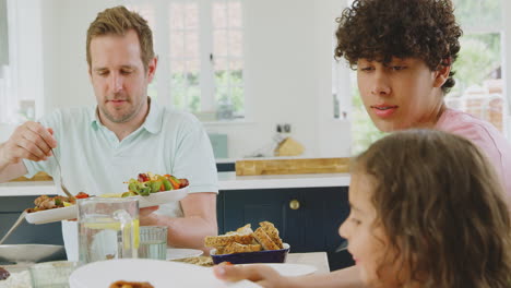 Vater-Dient-Als-Multiethnische-Familie,-Die-Zu-Hause-In-Der-Küche-Am-Tisch-Sitzt-Und-Gemeinsam-Isst