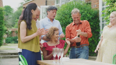 Gruppe-Reifer-Freunde-Bei-Einer-Überraschungs-Geburtstagsfeier-Für-Eine-Frau-Im-Garten-Mit-Kuchen-Und-Champagner